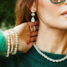 Load image into Gallery viewer, Close-up of a person wearing gold jewelry including earrings, rings, and bracelets against a blurred background.